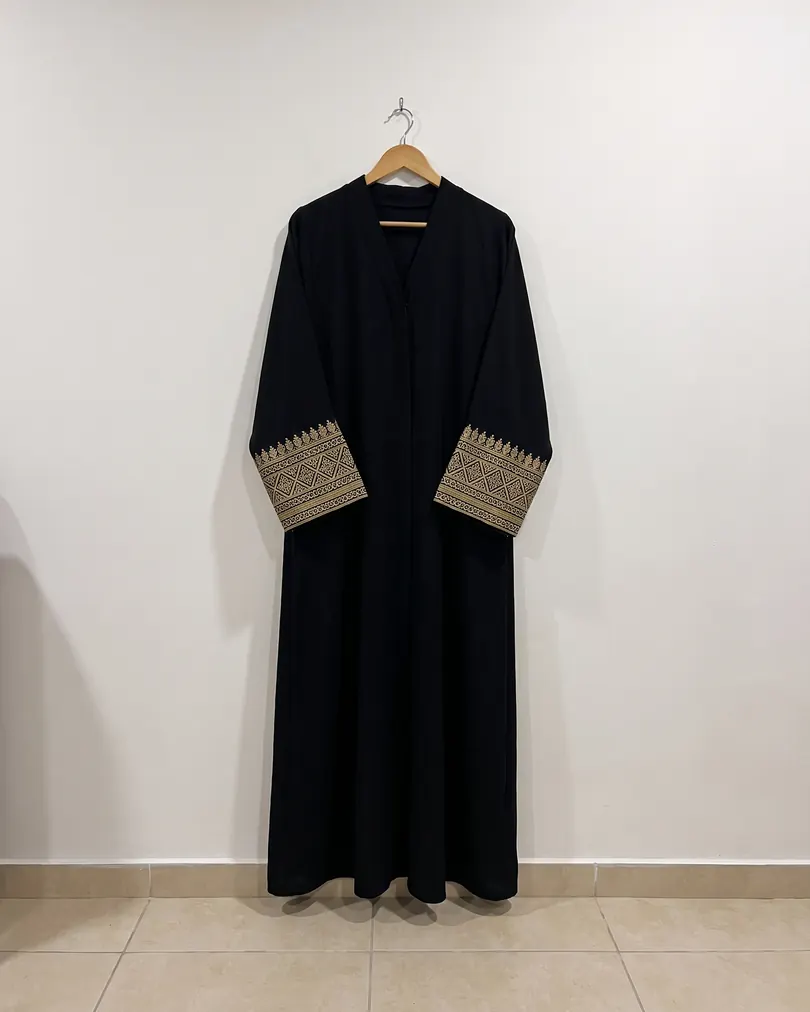 Black abaya with gold embroidered cuffs hanging on a white wall, shot on a phone