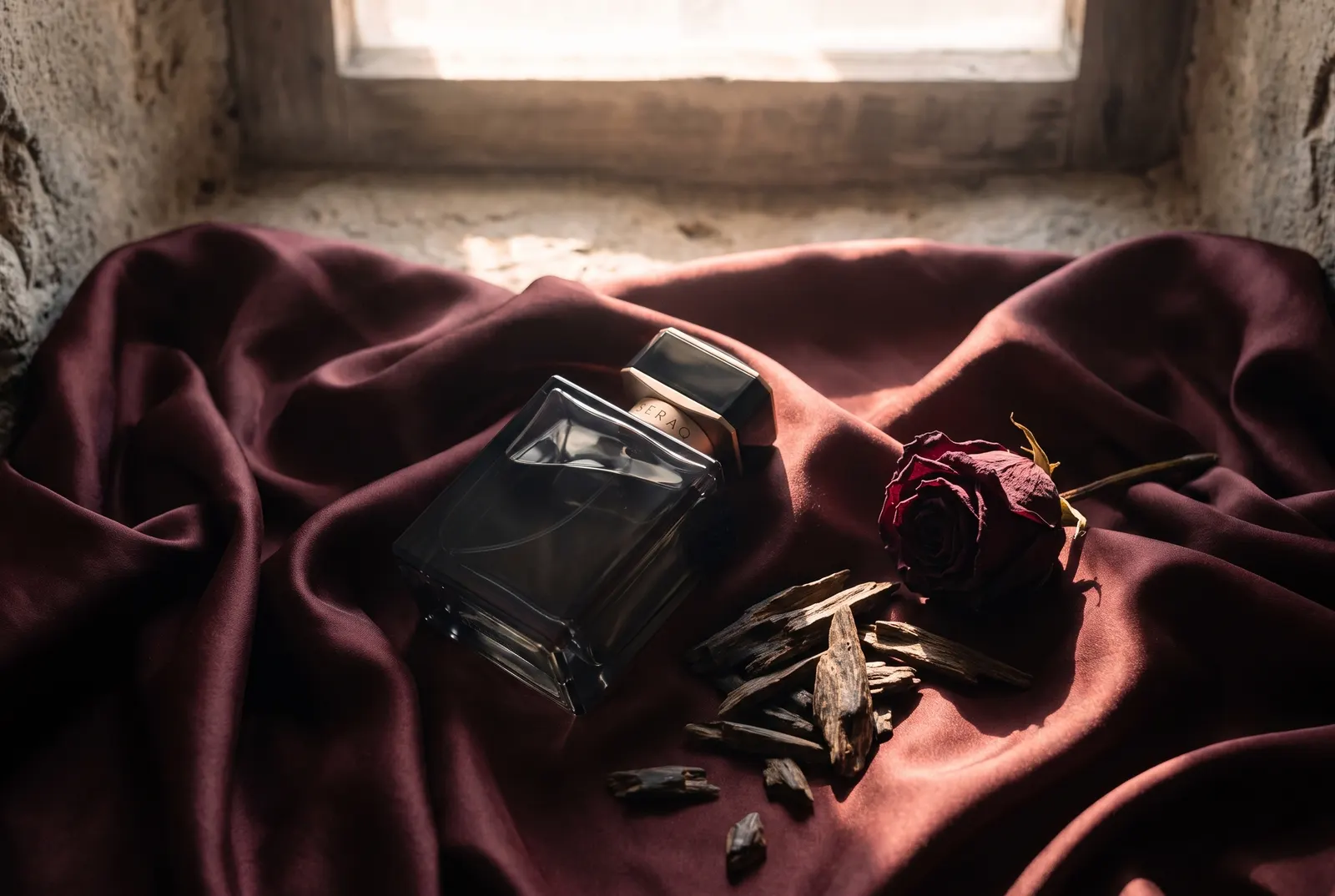 Perfume bottle on oxblood silk with oud wood and a dried rose