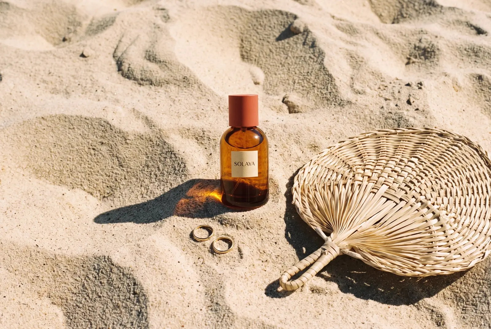 Body oil bottle in rippled sand with gold rings and a palm fan
